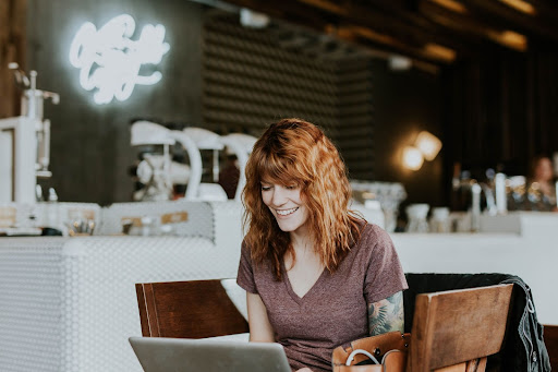 Red-Headed Woman Smiling Looking At Her Laptop In A Cafe