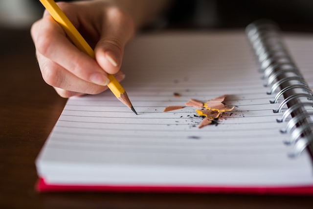 A hand holding a pencil to a blank notepad, with pencil shavings on it